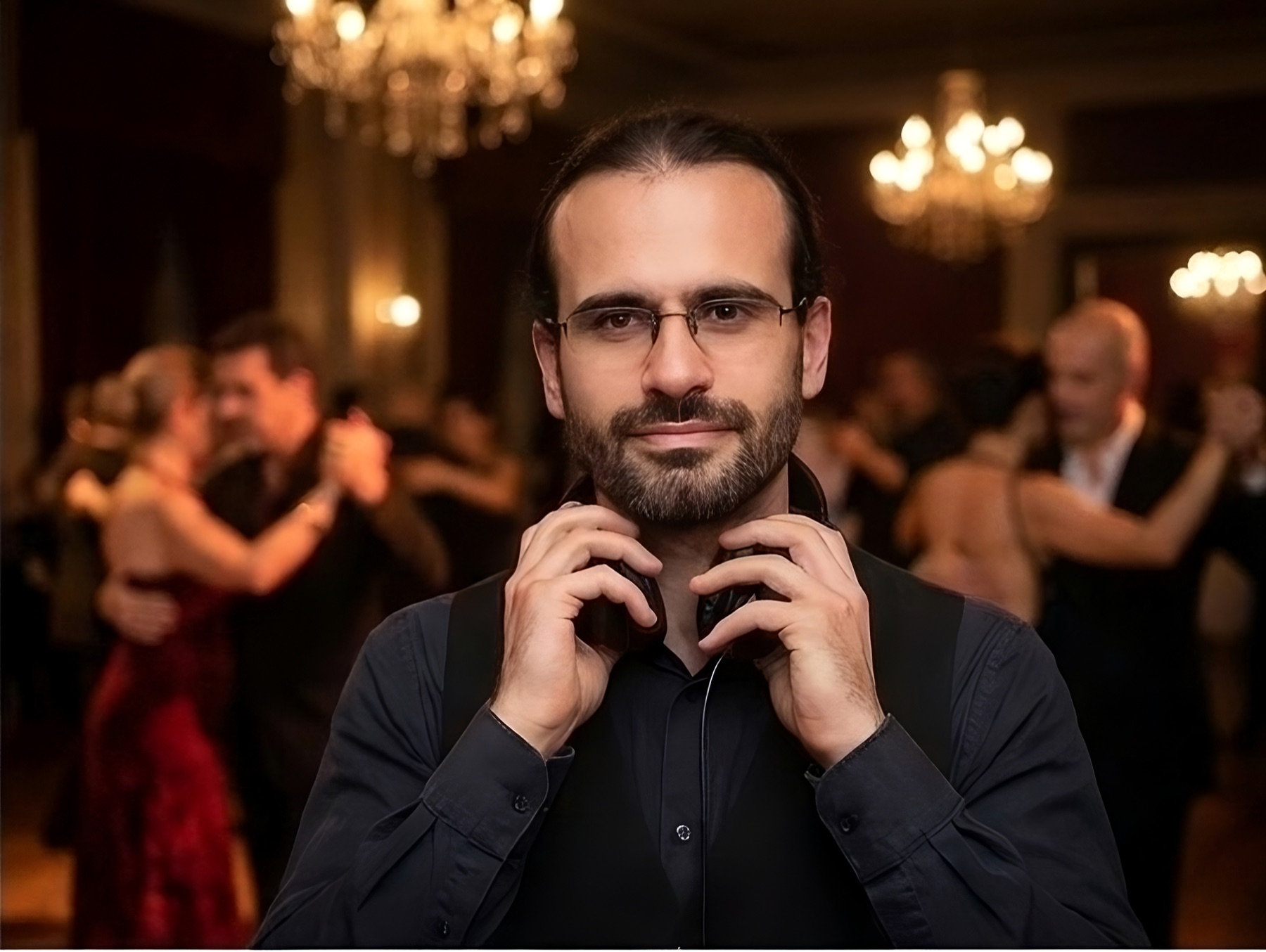 el-eje DJing at a tango milonga, headphones around his neck, dancers behind